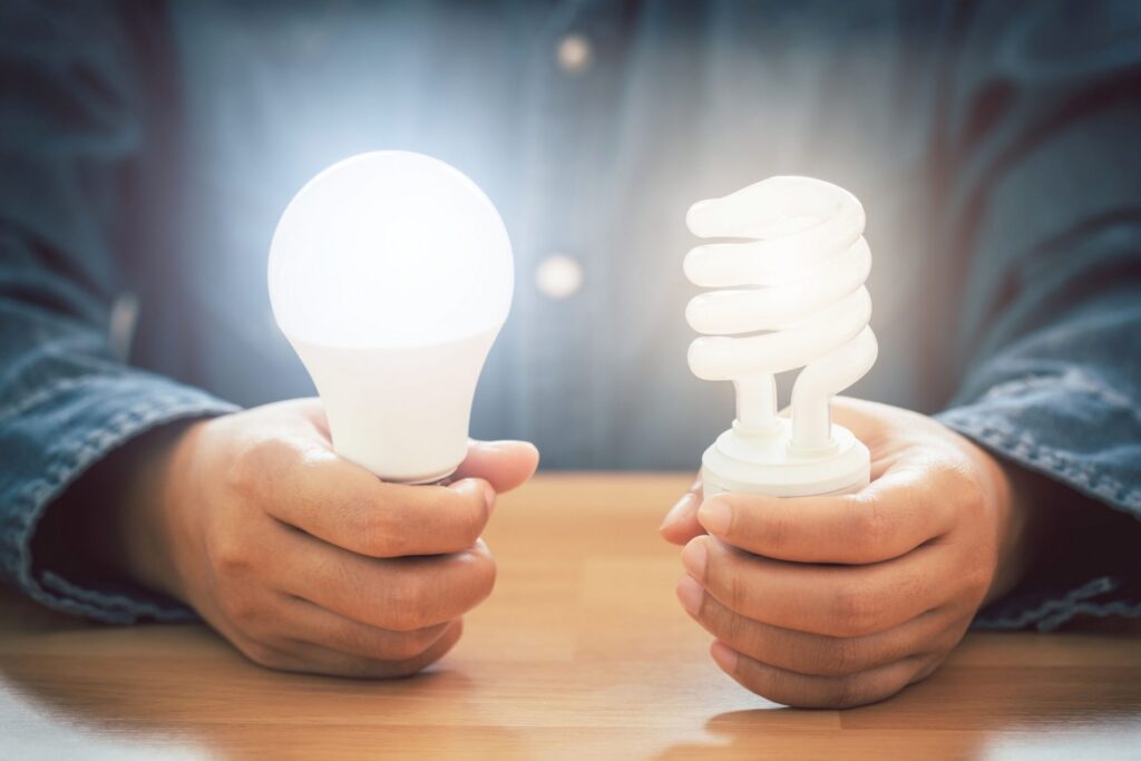 man holding two light bulbs that are lit up. the one on the left is an LED bulb and the one on the right is a spiral bulb