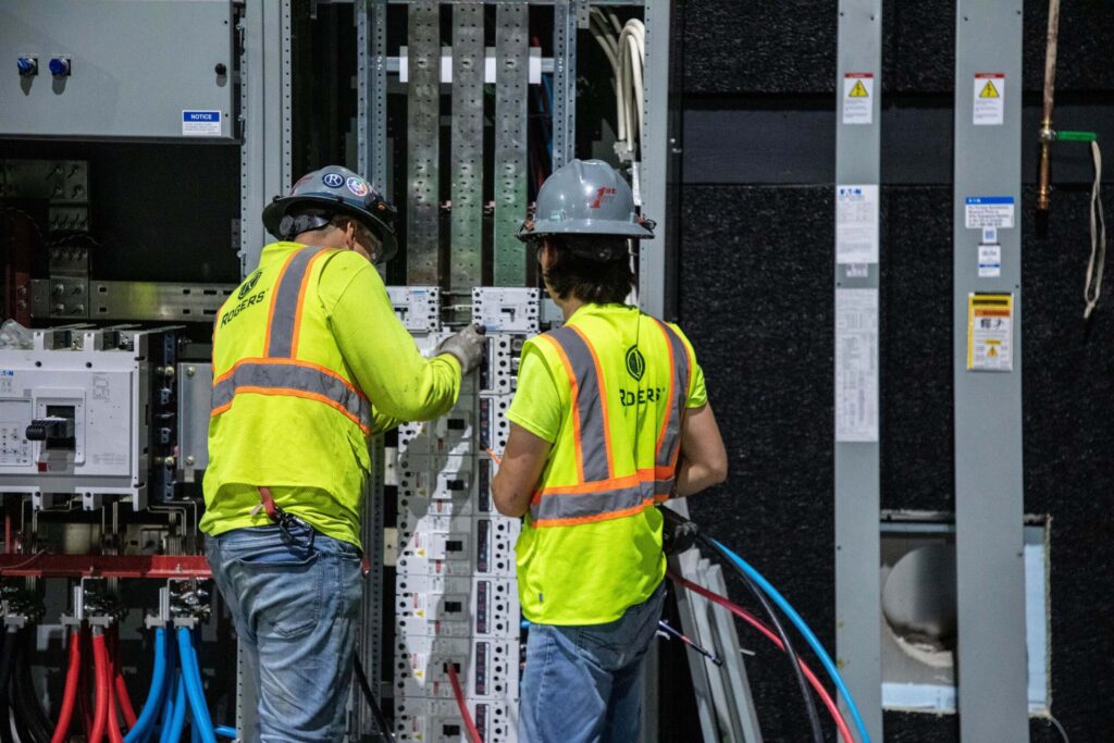 backside of two Rogers Electric employees in yellow safety vests with hard hats looking at electrical panel