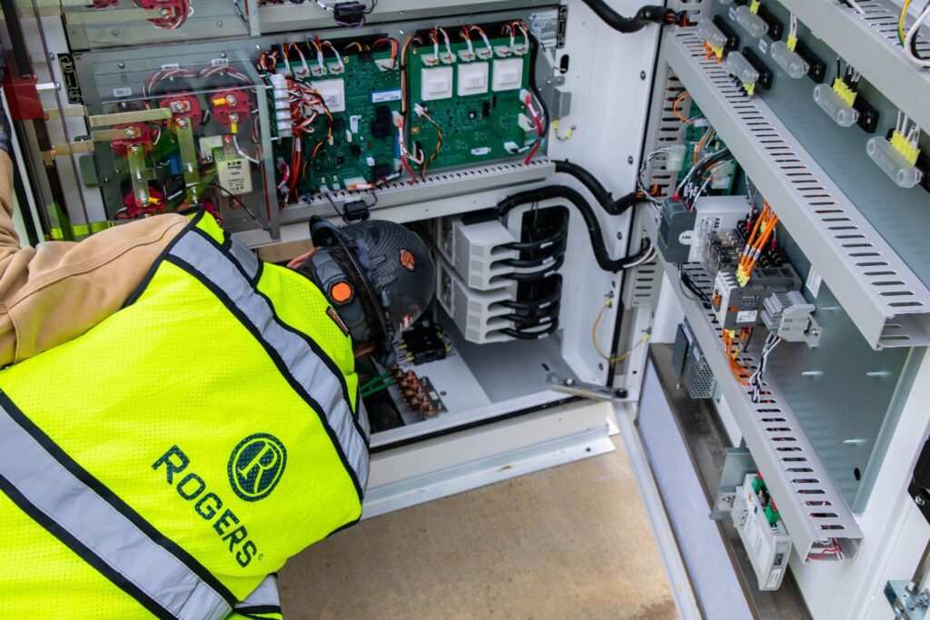 backside of electrician with Rogers safety vest and hard hat leaning over and looking into electrical panel