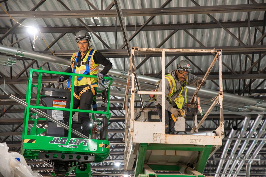 two electricians in two different scissor lifts, one green, one white- they are wearing yellow safety vests and hard hats while being harnessed into the lifts