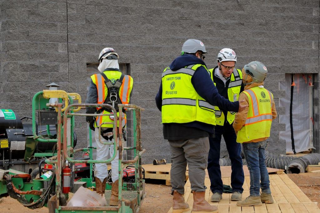 four electricians in yellow safety vests and hard hats on a construction site- one is turned away and two are talking to a shorter electrician