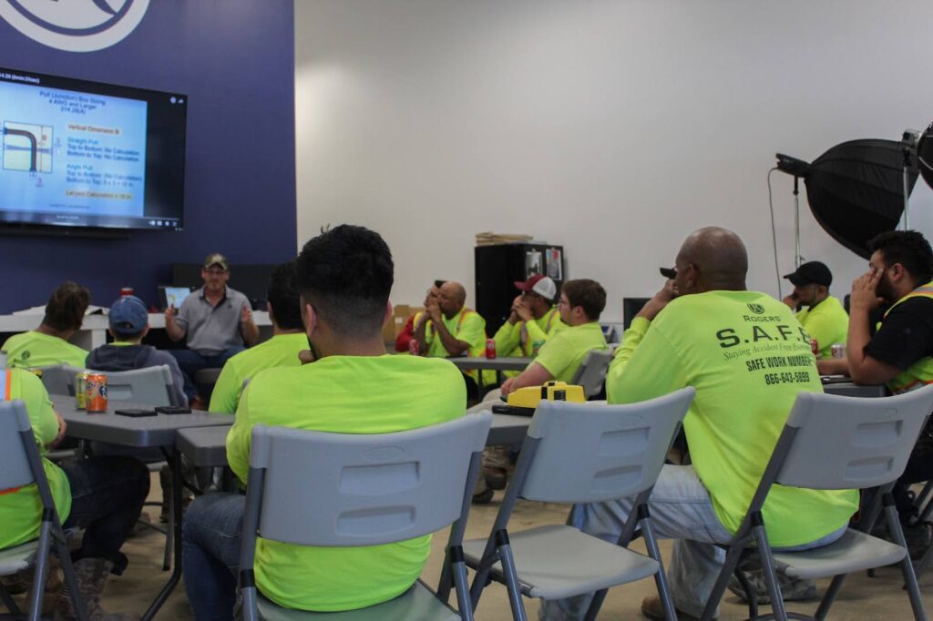 a large room of electricians in chairs facing a screen all wearing bright yellow long sleeve shirts in a training class