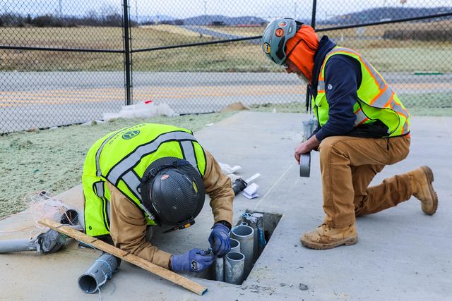 two electricians working on piping under the ground. One is bending over working on piping and the other is on his knees with duct tape ready to go