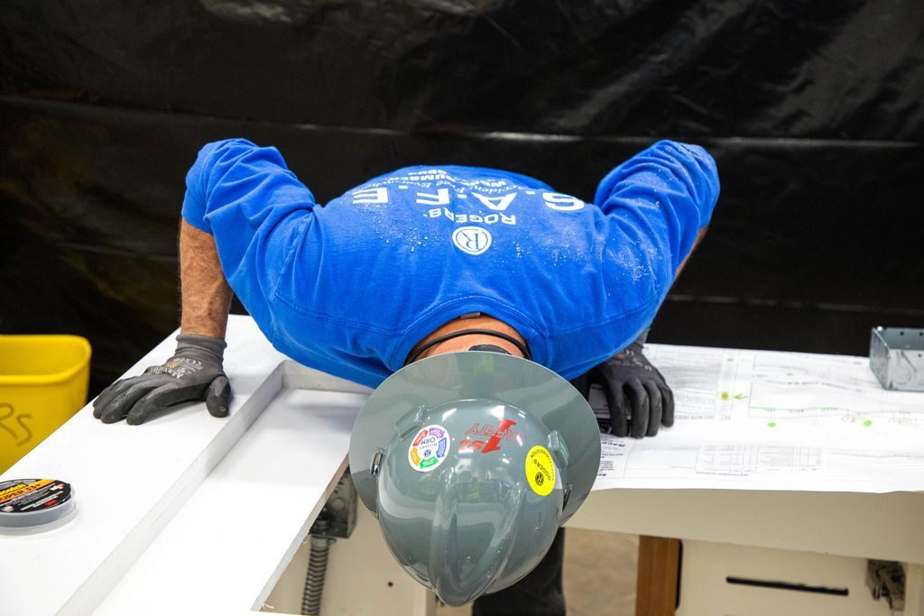 electrician in blue long sleeve shirt bending over a counter. He is wearing gloves and a gray hard hat with stickers on it
