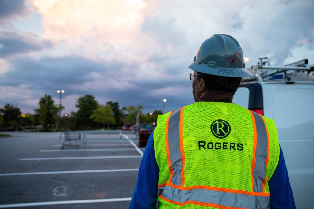 back of an electrician in a parking lot with a Rogers safety vest and hard hat and a pretty sunset