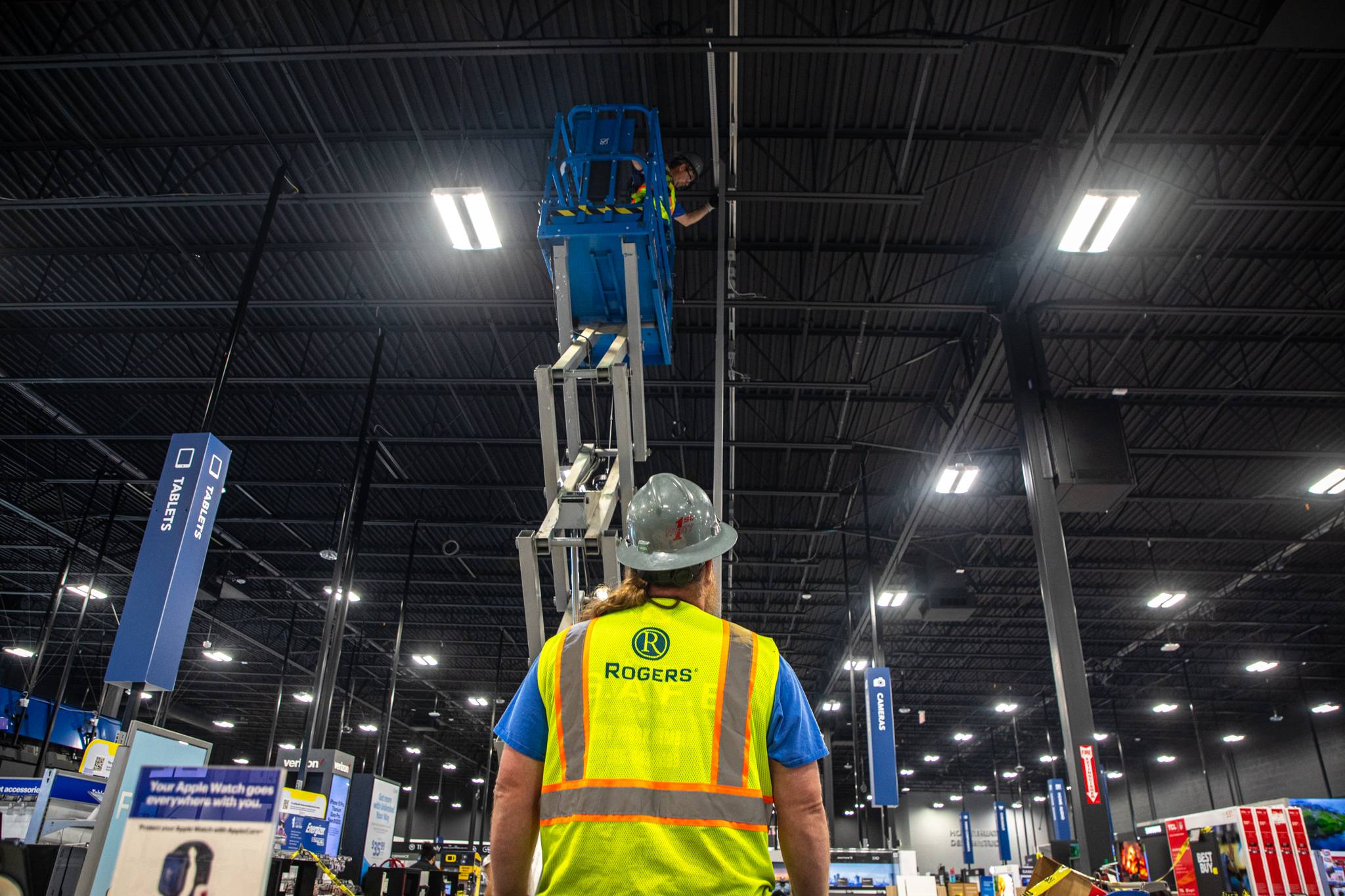 Electrician looking up at an electrician in a scissor lift inside of a Best Buy with Rogers showing on his safety vest