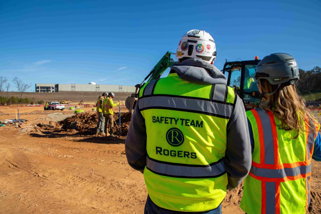 Two electrical construction workers, a man and a woman looking off observing the jobsite. One worker is wearing a safety team vest.