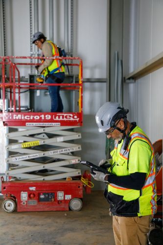 Two commercial electricians working on a job site. One is on a scissor lift, and the other is looking at blueprints on a tablet.