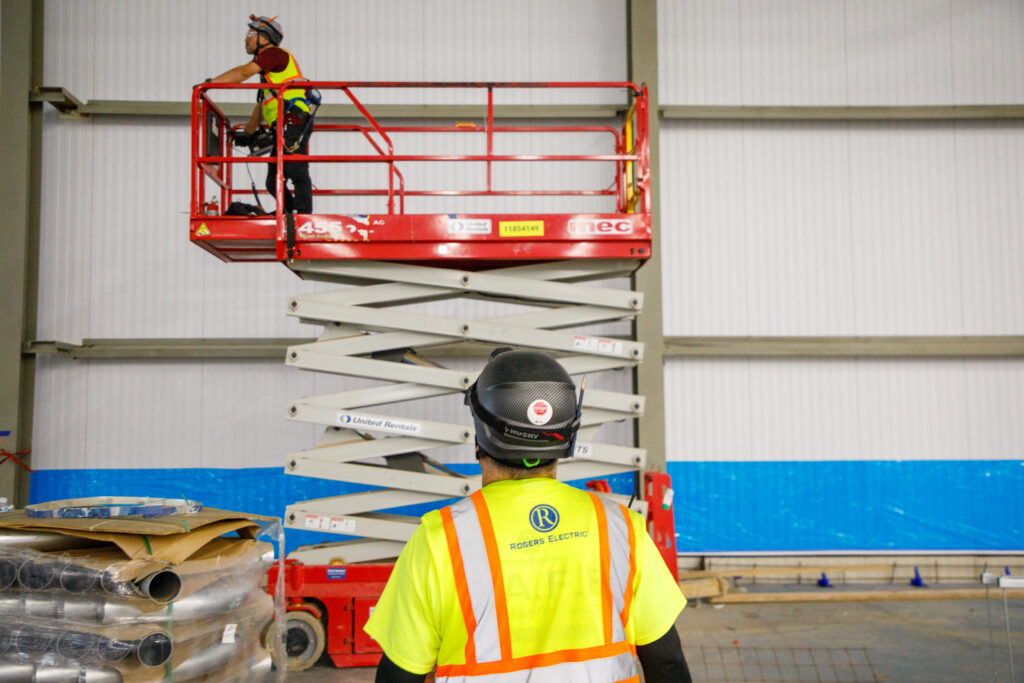 Two commercial electricians in a warehouse. One is on a scissor lift and the other is looking at the scene.