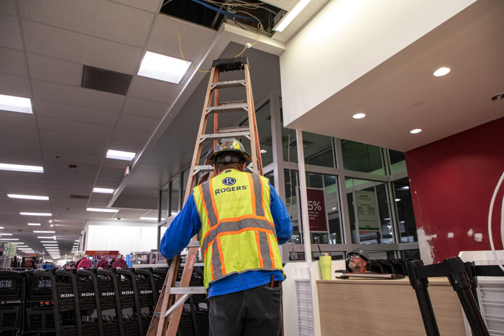 Rogers Electric technician on a ladder working above a retail ceiling, upgrading electrical systems inside a commercial space.